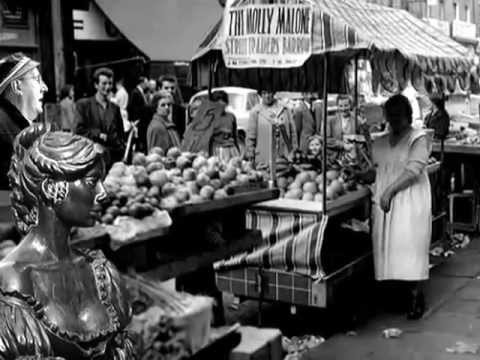 A crowded outdoor market scene with fruit and vegetable stalls, a striped canopy, and a statue of a woman in the foreground. Shoppers browse produce, and a woman stands near a stall, partially in shadow.