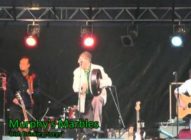 A band performs on stage under colored lights. One musician in the center stands with a drum, while others play guitars. The text Murphys Marbles Irish Drinking Songs appears at the bottom left.