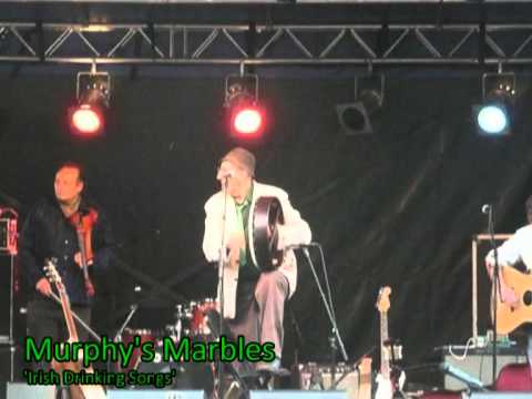 A band performs on stage under colored lights. One musician in the center stands with a drum, while others play guitars. The text Murphys Marbles Irish Drinking Songs appears at the bottom left.