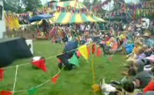 A colorful outdoor festival with people sitting on grass, surrounded by vibrant triangular pennant banners. A large yellow-and-green striped tent and festive decorations fill the background.