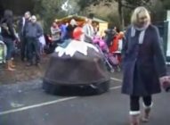 A person dressed in a large Christmas pudding costume stands among a crowd at an outdoor event; people in coats and scarves walk by, and market stalls are visible in the background.