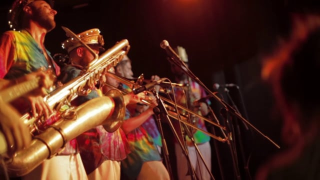 A brass band wearing colorful, tie-dye shirts and hats plays trombones and saxophones on stage under warm lighting, with microphones set up in front of them.