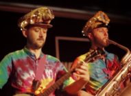 Two musicians wearing colorful tie-dye shirts and shiny gold hats perform on stage; one plays a banjo, and the other plays a large brass instrument, possibly a saxophone.