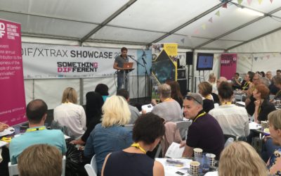 A man speaks at a podium in a large white tent to an audience seated at tables, with banners, presentation screens, and event materials visible around the room.