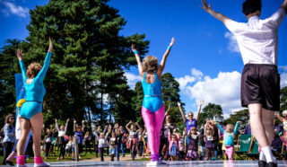Three performers in colorful workout outfits lead a large outdoor group in an exercise routine, with everyone raising their arms under bright blue skies and surrounded by trees.