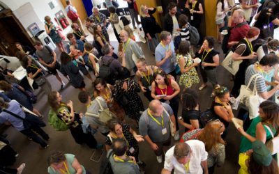 Large group of people wearing conference name tags gather and mingle in a bright indoor setting, conversing, holding drinks, and interacting in small groups. Photographed from above.