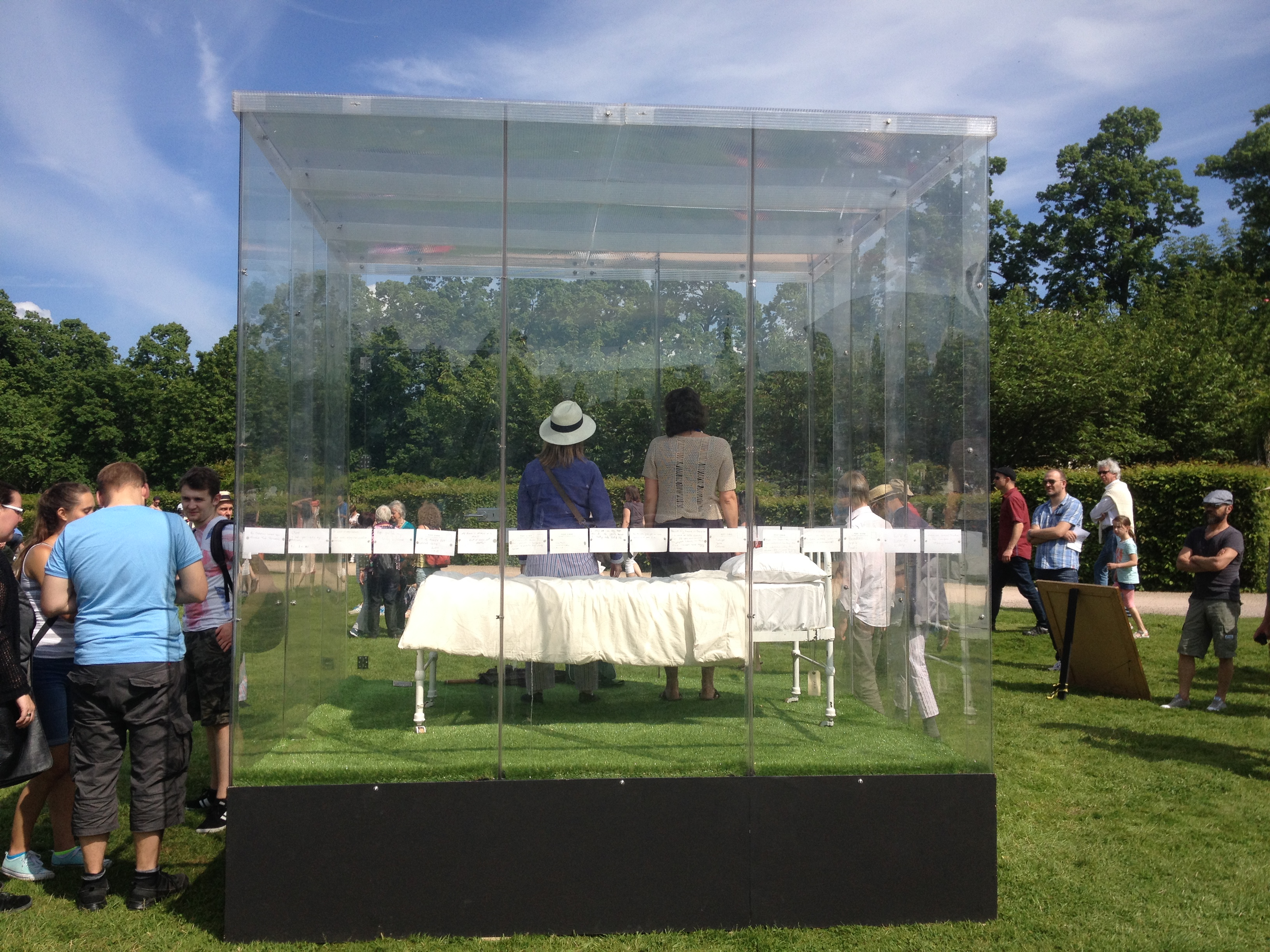 A group of people gather outdoors around a transparent glass enclosure containing a white-covered table, with two people inside. The scene is set on a grassy lawn under a blue sky with scattered clouds.