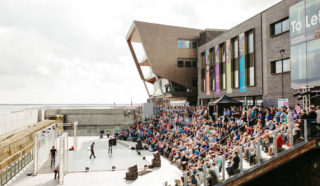 A large crowd sits on outdoor steps watching performers on a stage near a waterfront building with modern architecture, colorful banners, and glass windows under a partly cloudy sky.