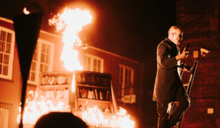A person in a dark coat stands on a ladder next to a large bookshelf structure engulfed in flames, with fire dramatically shooting upwards, set outdoors at night in front of a brick building.