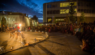 A performer sits on stage under bright lights in an outdoor plaza at dusk, while a large audience watches. Surrounding buildings are visible, including a “Bingo” sign, with the sky turning dark blue above.