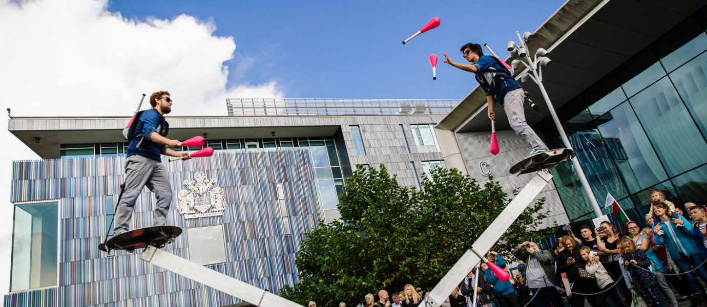Two performers balance on opposite ends of a large seesaw while juggling clubs, entertaining a crowd gathered outside a modern building on a sunny day.
