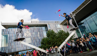 Two performers balance on opposite ends of a large seesaw while juggling clubs, entertaining a crowd gathered outside a modern building on a sunny day.