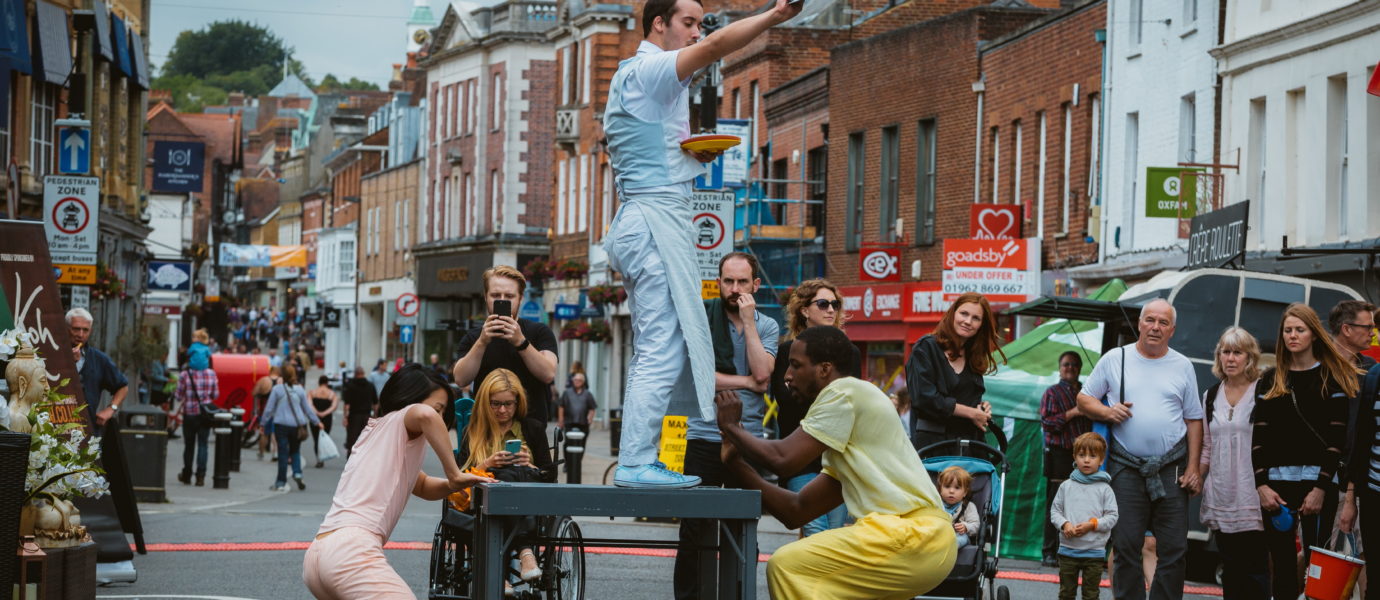 Three performers in colorful outfits balance on and around a table in a lively street, entertaining a diverse crowd of onlookers, including children and people in wheelchairs. Buildings and shops line the background.
