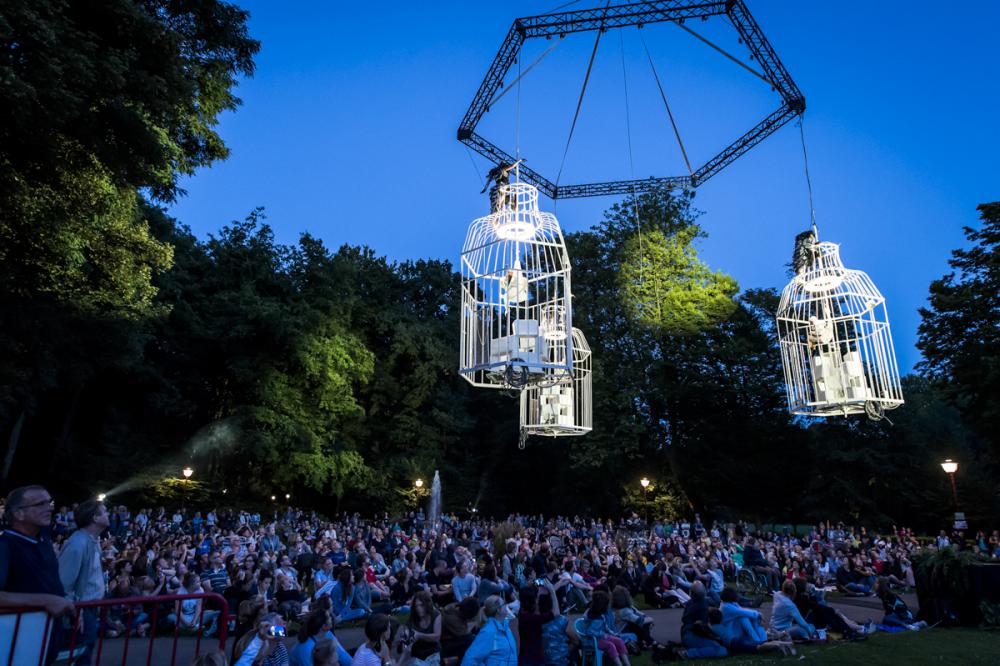 A large crowd gathers outdoors at dusk, watching performers in giant birdcages suspended high above the ground among tall trees, illuminated by spotlights.