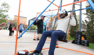 A man sitting on a swing set wears a virtual reality headset, holding the chains, while another smiling man stands nearby outdoors. People and equipment are visible in the background.