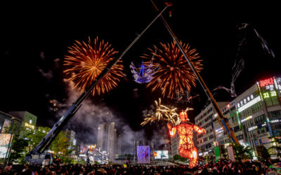 A vibrant night scene shows fireworks bursting over a crowded city square, with large illuminated puppets and cranes in the center, surrounded by tall buildings with bright signs.