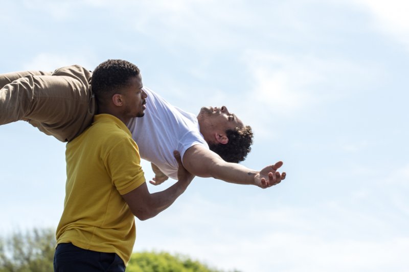 Two men are outdoors; one in a yellow shirt lifts the other, who is wearing a white shirt, with arms outstretched, creating a dramatic pose against a blue sky background.