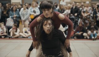 Two women perform a dance in an outdoor public space, with one woman crouched and the other perched on her back. A large, diverse crowd watches the performance in the background.