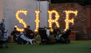 People sit and relax on grass near several pushchairs in front of large illuminated letters spelling SIRF against a brick and white wall, with fairy lights above, suggesting a festive outdoor event.