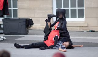 Two performers in costume act out a dramatic scene on an outdoor stage. One wears a red mask and black veil, posing with a leg raised, whilst the other lies on the ground in casual clothes, appearing motionless.