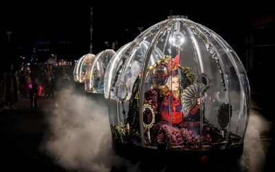 A woman in an elaborate red costume and mask sits inside a transparent, ornate carriage shaped like a dome, surrounded by fog, during a nighttime parade with similar carriages in the background.