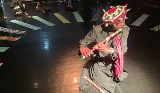 A person in a gray outfit and colorful, beaded hat plays a flute while kneeling on a dimly lit stage, with patterned fabric strips on the floor and several people seated in the background.