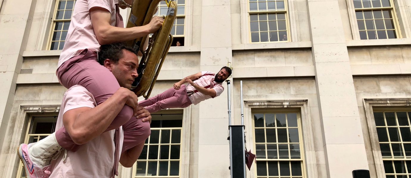 Three performers in matching pink outfits: one balancing another on his shoulders, while the one on top plays a tuba. A third performer is mid-air above a trampoline, in front of a classical building with large windows.