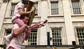 Three performers in matching pink outfits: one balancing another on his shoulders, while the one on top plays a tuba. A third performer is mid-air above a trampoline, in front of a classical building with large windows.