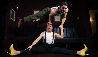 Two men perform an acrobatic act indoors; one does a split on the floor wearing yellow high heels, while balancing the other man on his head with one hand. Both wear suspenders and white shirts. The room is dimly lit.