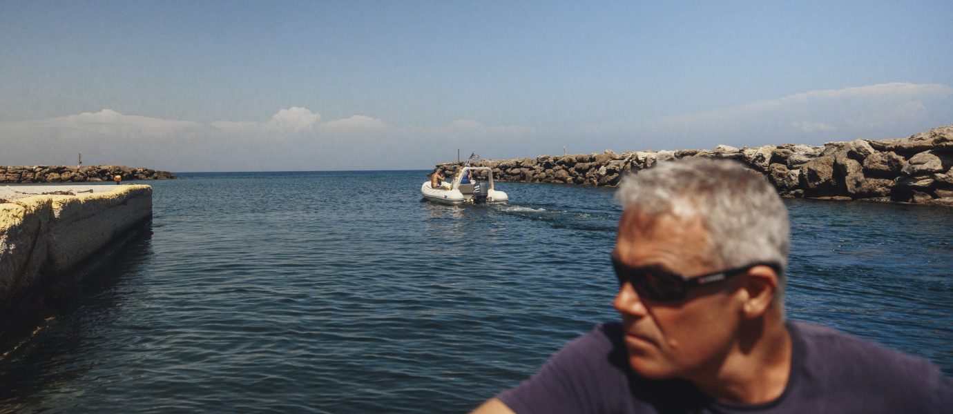 A man in sunglasses sits on a boat in the foreground, looking to the side. In the background, a small motorboat approaches through a rocky channel under a clear blue sky.