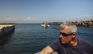 A man in sunglasses sits on a boat in the foreground, looking to the side. In the background, a small motorboat approaches through a rocky channel under a clear blue sky.