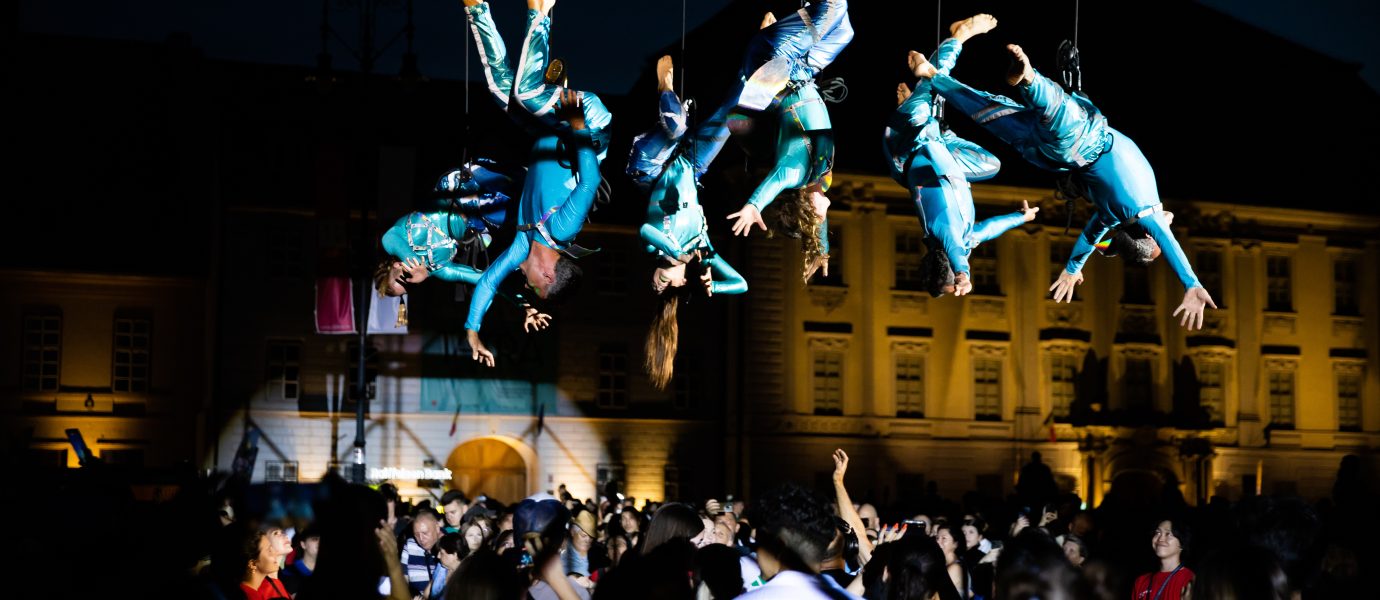 Performers in blue costumes hang upside down on wires above a large crowd at night, with a historic building illuminated in the background. The crowd watches the aerial show attentively.