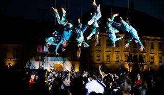 Performers in blue costumes hang upside down on wires above a large crowd at night, with a historic building illuminated in the background. The crowd watches the aerial show attentively.