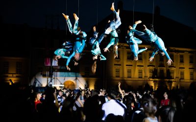 Performers in blue costumes hang upside down on wires above a large crowd at night, with a historic building illuminated in the background. The crowd watches the aerial show attentively.