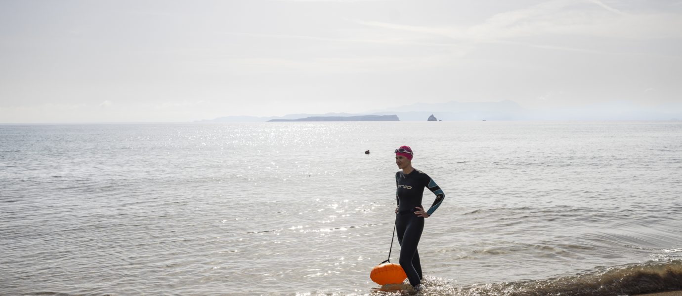A person in a wetsuit and swim cap stands on a sandy beach at the water's edge, holding an orange float. The sea is calm, and distant islands are visible on the horizon under a hazy sky.
