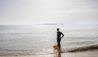 A person in a wetsuit and swim cap stands on a sandy beach at the water's edge, holding an orange float. The sea is calm, and distant islands are visible on the horizon under a hazy sky.
