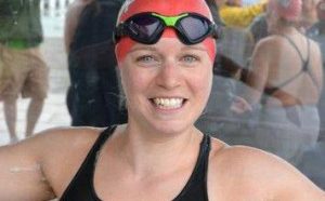 A smiling swimmer in a black swimming costume, red swim cap, and goggles stands with arms raised. Other swimmers in similar kit are visible in the background. The scene appears to be at an outdoor swimming event.