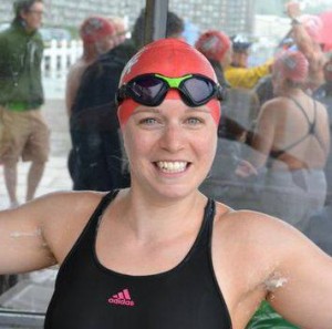 A smiling swimmer in a black swimming costume, red swim cap, and goggles stands with arms raised. Other swimmers in similar kit are visible in the background. The scene appears to be at an outdoor swimming event.