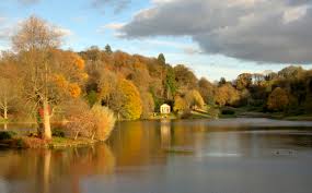 A tranquil lake surrounded by trees with autumn foliage, reflecting shades of yellow, orange, and brown. A small white structure sits near the water's edge under a partly cloudy sky.